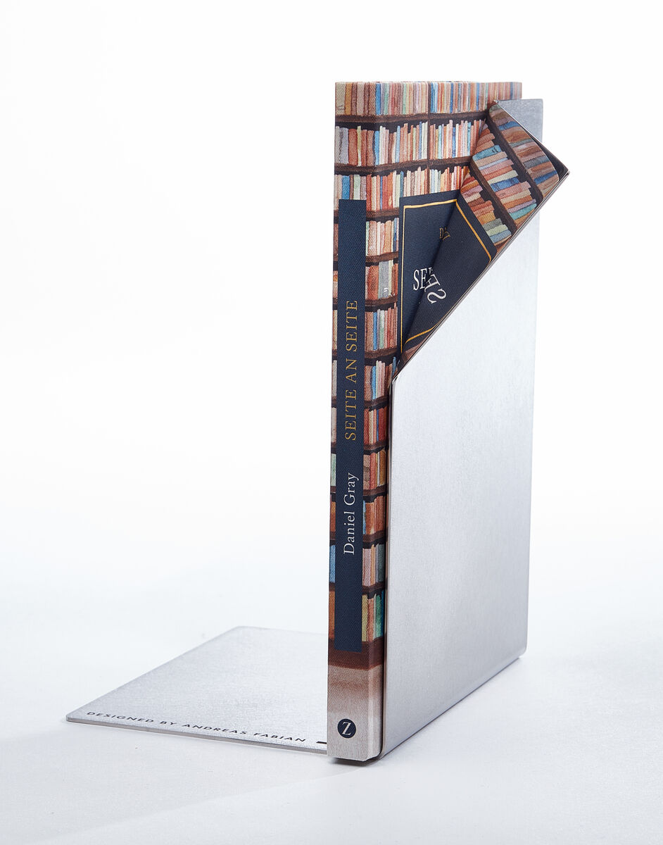 Eine Buchstütze aus Metall, die im Bild eine aufgeschlagene Buchseite imitiert, hält Bücher in einem Regal. Der Hintergrund ist weiß und minimalistisch gehalten.