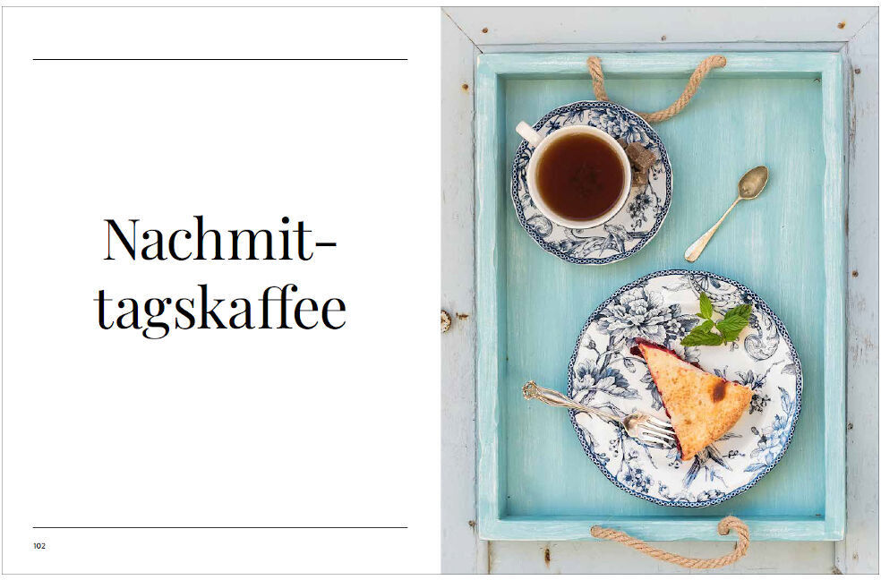 Aufgeschlagenes Kochbuch mit dem Titel 'Sonntagsessen', geöffnete Seite zeigt das Wort 'Nachmittagskaffee' links und rechts eine Tasse Kaffee, ein Kuchenteller und ein Löffel auf einem hellblauen Holztablett.