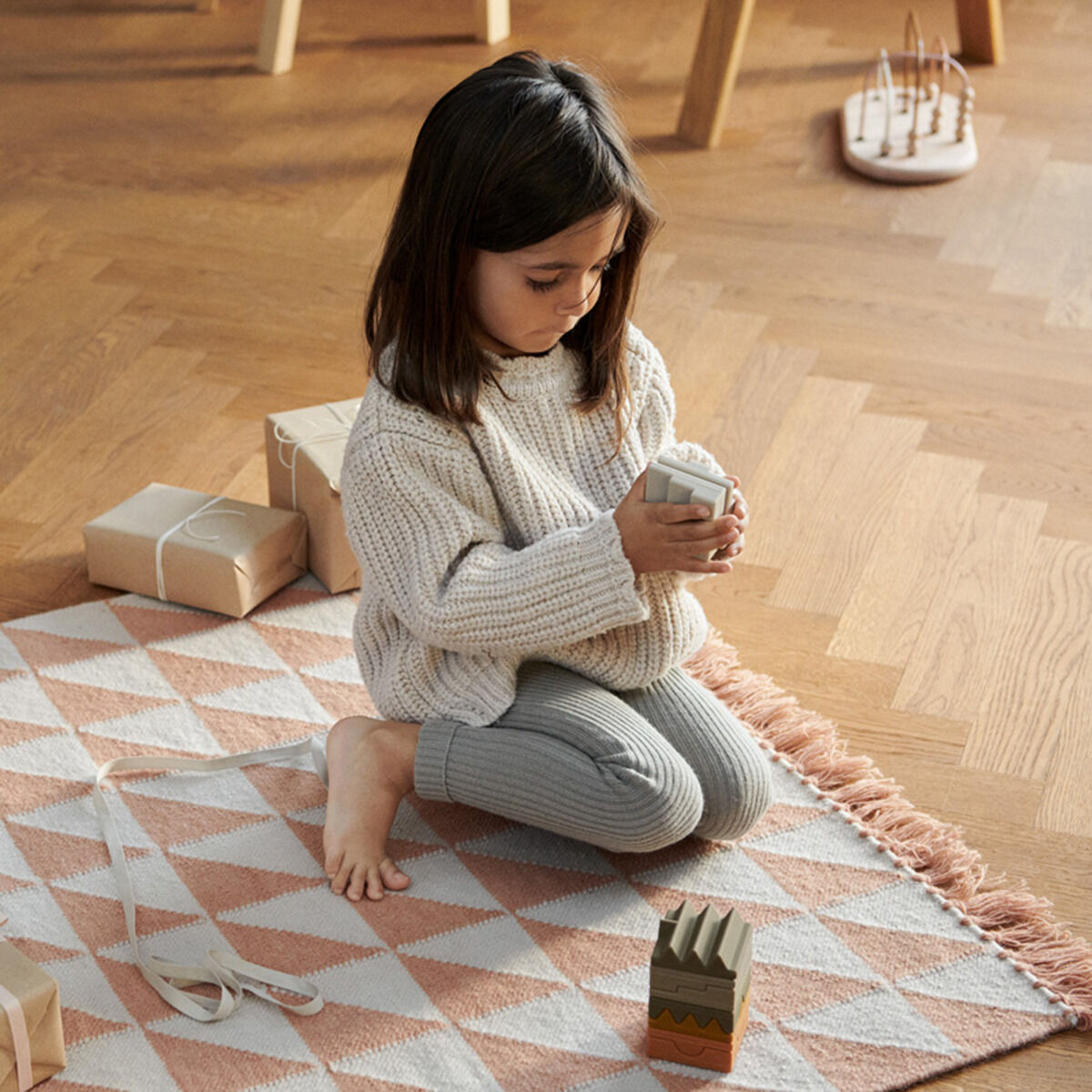 Ein Kind sitzt auf einem gemusterten Teppich und spielt mit einem Stapelturm aus Holzringe. Es hat graue Leggings und einen weißen Pullover an. Im Hintergrund sind ein kleines Holztischchen und ein weiteres Spielzeug auf dem Holzboden zu sehen.