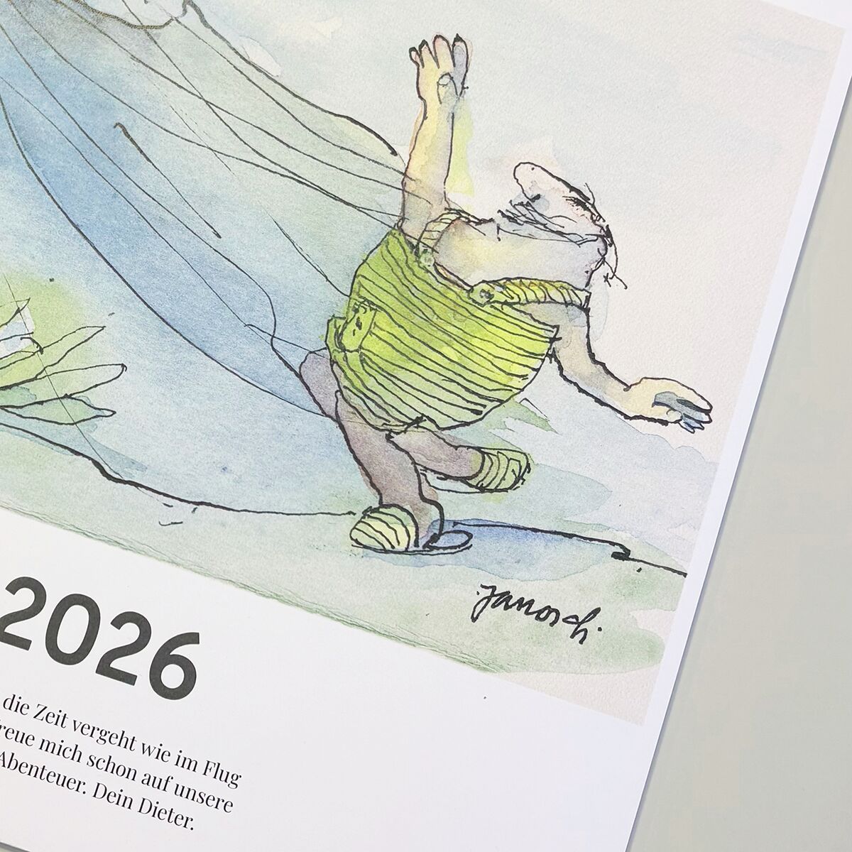 Titelbild eines Wandkalenders für das Jahr 2026 mit einer humorvollen Illustration einer Tierfigur im Comicstil, die ein grünes gestreiftes Kleid trägt. Der Kalender trägt die Aufschrift 'Wondrak-Kalender 2026' und ist auf eine helle, moderne Oberfläche gelegt.