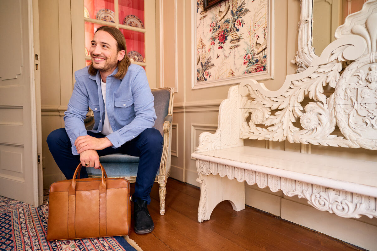 Ein Mann sitzt in einem eleganten Raum auf einem Stuhl und hält eine hellbraune Ledertasche »Harvey«. Er trägt eine blaue Hemdjacke und eine dunkelblaue Hose.