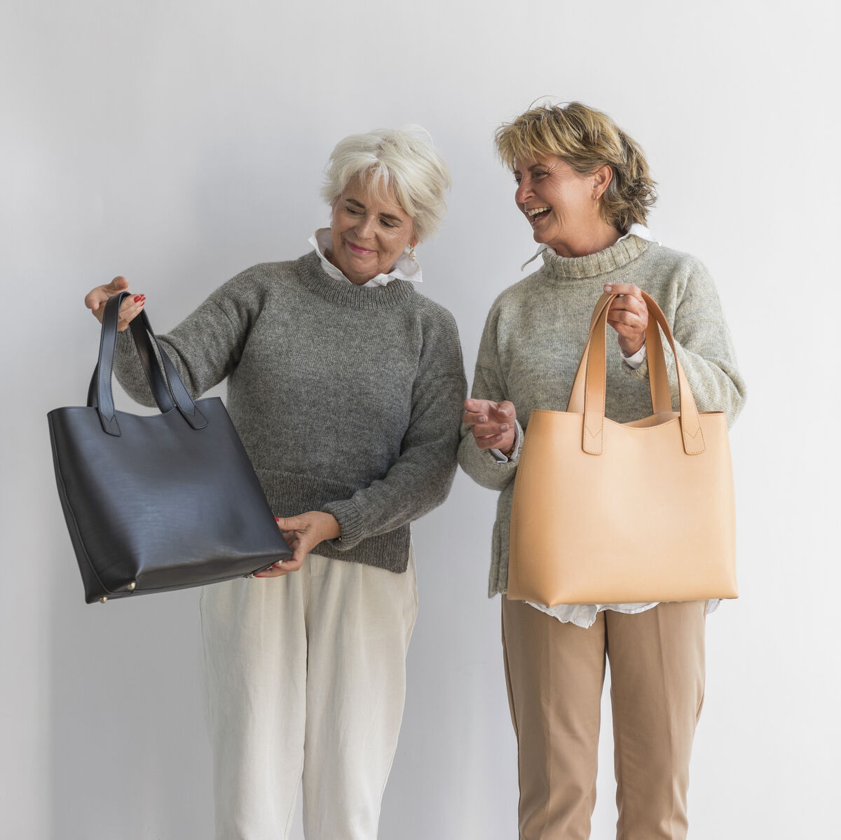 Zwei Frauen halten jeweils eine große Tasche aus Kernleder vor einem weißen Hintergrund. Eine Tasche ist schwarz und die andere ist hellbraun. Beide Frauen tragen graue Pullover.