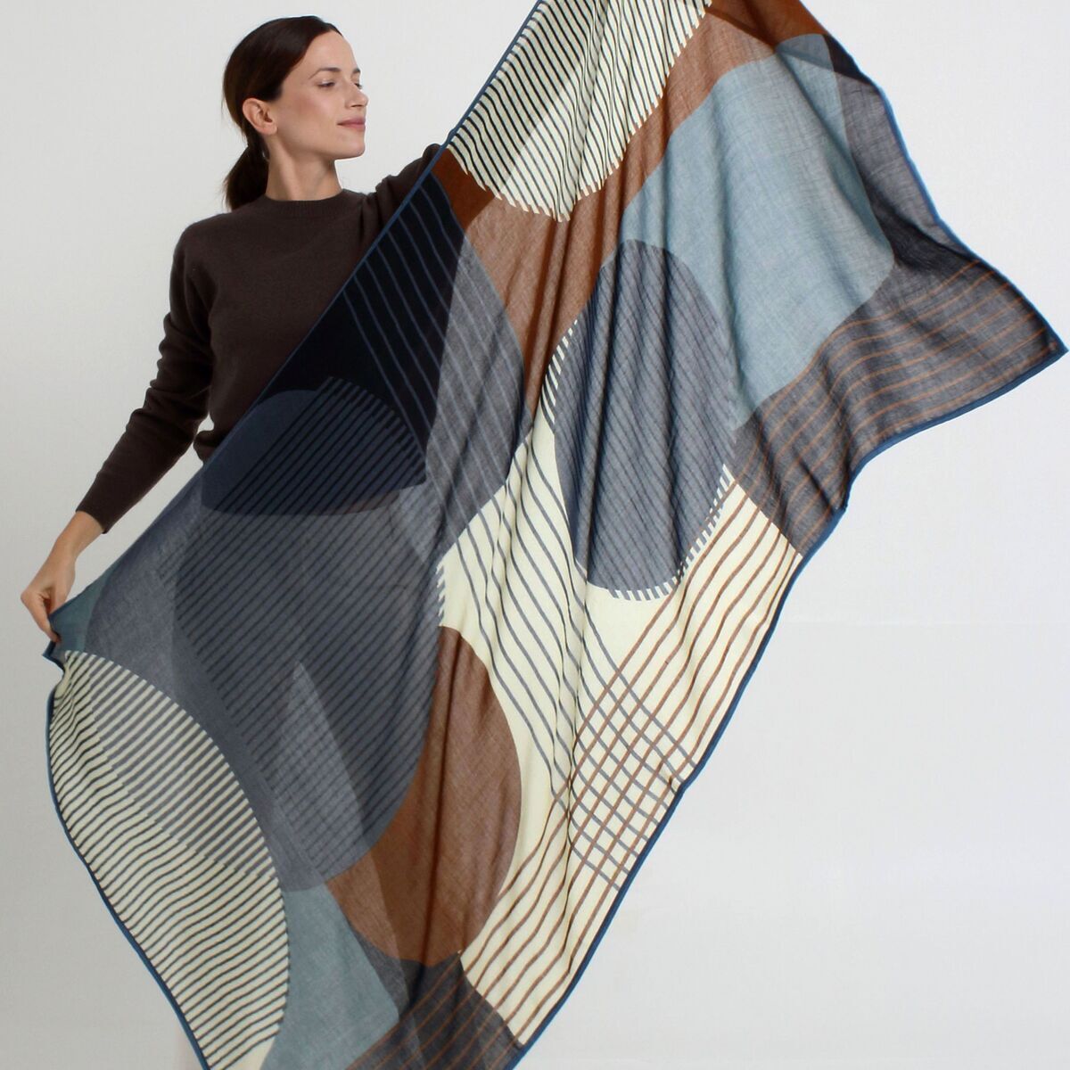 Eine Frau hält den großen Wollschal »Alma« der Marke Shalls Barcelona aus 100% Schurwolle. Der Schal zeigt ein geometrisches Muster in Blau-, Braun-, Creme- und Beigetönen mit Streifen und Kreisen. Der Schal wirkt leicht und hochwertig verarbeitet.
