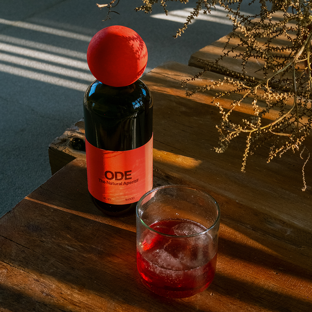 Flasche mit rotem Etikett und rotem runden Verschluss neben Glas mit rotem Getränk auf Holztisch, Sonnenlicht und Pflanzenzweig im Hintergrund