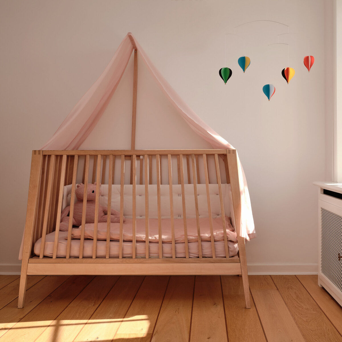 Ein Babyzimmer mit einem Holzgitterbett, das mit rosa Bettwäsche ausgestattet ist. Darüber hängt ein Mobile mit fünf bunten Heißluftballons. Links im Bett liegt ein Kuscheltier. Rechts steht ein weißer Schrank unter einem Fenster mit Sonnenlicht.