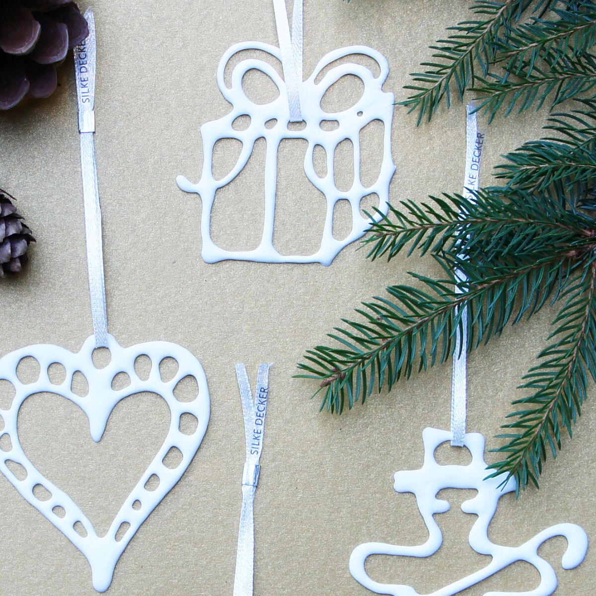 Vier filigrane Weihnachtsanhänger aus weißem Bonechina-Porzellan mit satinweißen Bändern liegen auf beigefarbenem Untergrund. Motive: Geschenkpaket, Herz, Stiefel und weiterer Anhänger. Eine grüne Tannenzweige und ein Pinienzapfen rahmen das Bild.