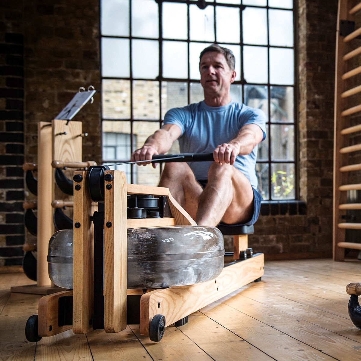 Ein Mann trainiert auf einem Waterrower-Rudergerät aus Eichenholz in einem stilvollen Raum mit Holzboden und Backsteinwänden.