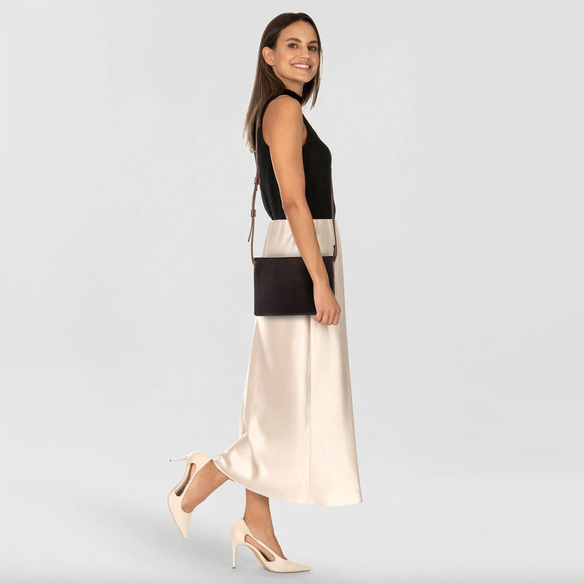 Eine Frau trägt eine elegante, dunkelbraune Kernledertasche »Sas« von Harold's Lederwaren als Umhängetasche. Das Outfit besteht aus schwarzem Top, cremefarbener Satinrock und cremefarbenen Pumps, vor hellem Hintergrund. Die Tasche wirkt schlicht und stilvoll.