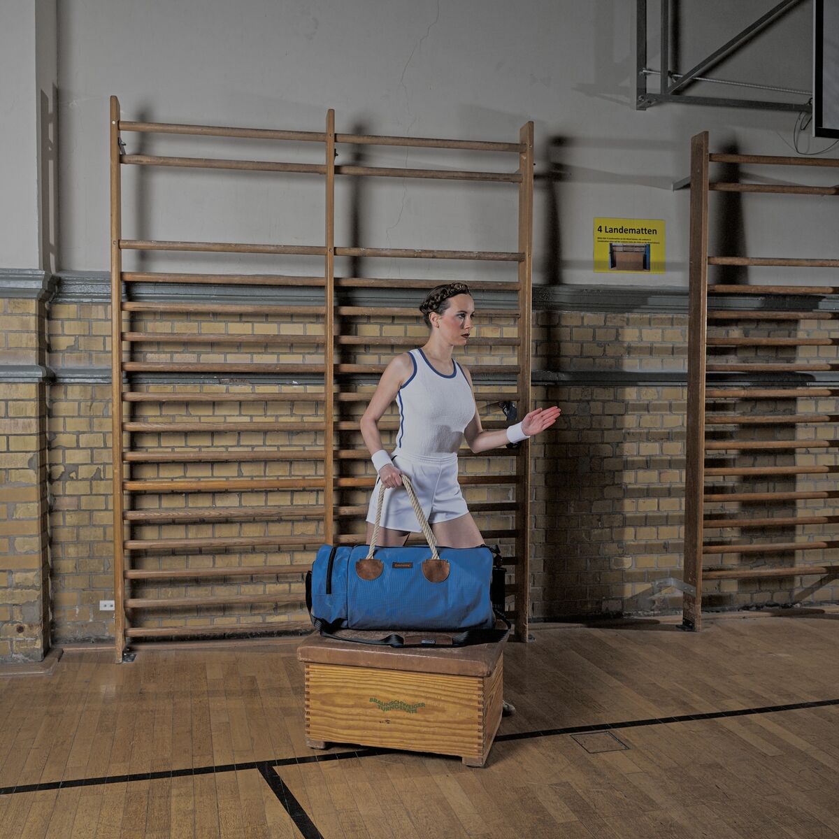 Eine Person in Sportkleidung hält eine blau-braune Sport- und Reisetasche »Rolle« in einer Turnhalle mit Holzboden und Sprossenwänden.