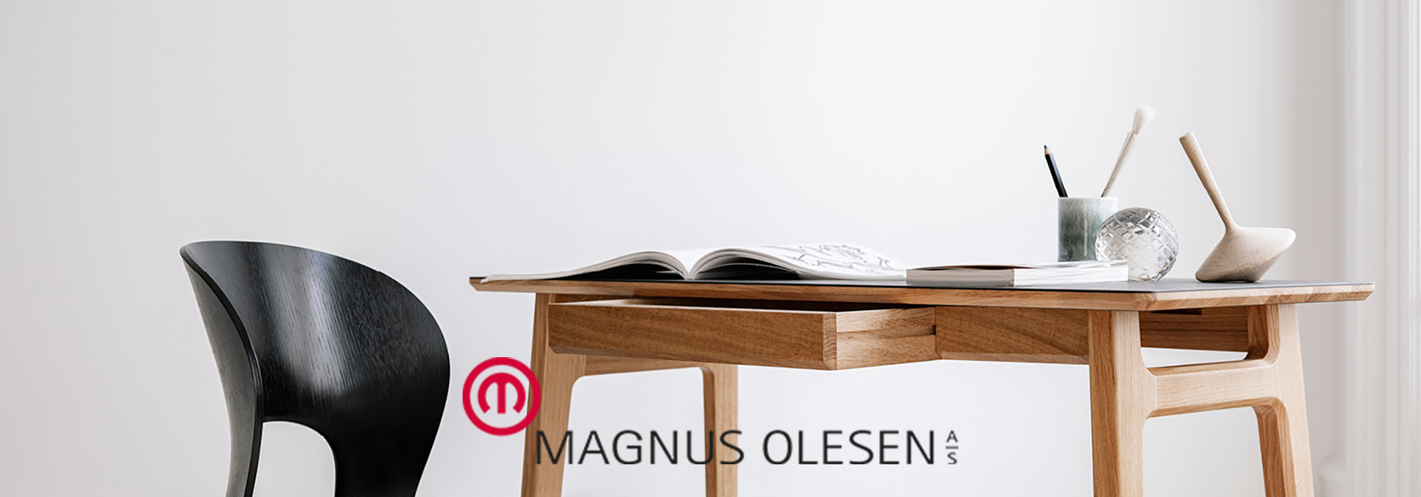 Ein stilvoller Holzschreibtisch mit einem aufgeschlagenen Magazin, daneben ein schlichter schwarzer Stuhl und Dekorationsartikel mit dem Schriftzug „Magnus Olesen“.