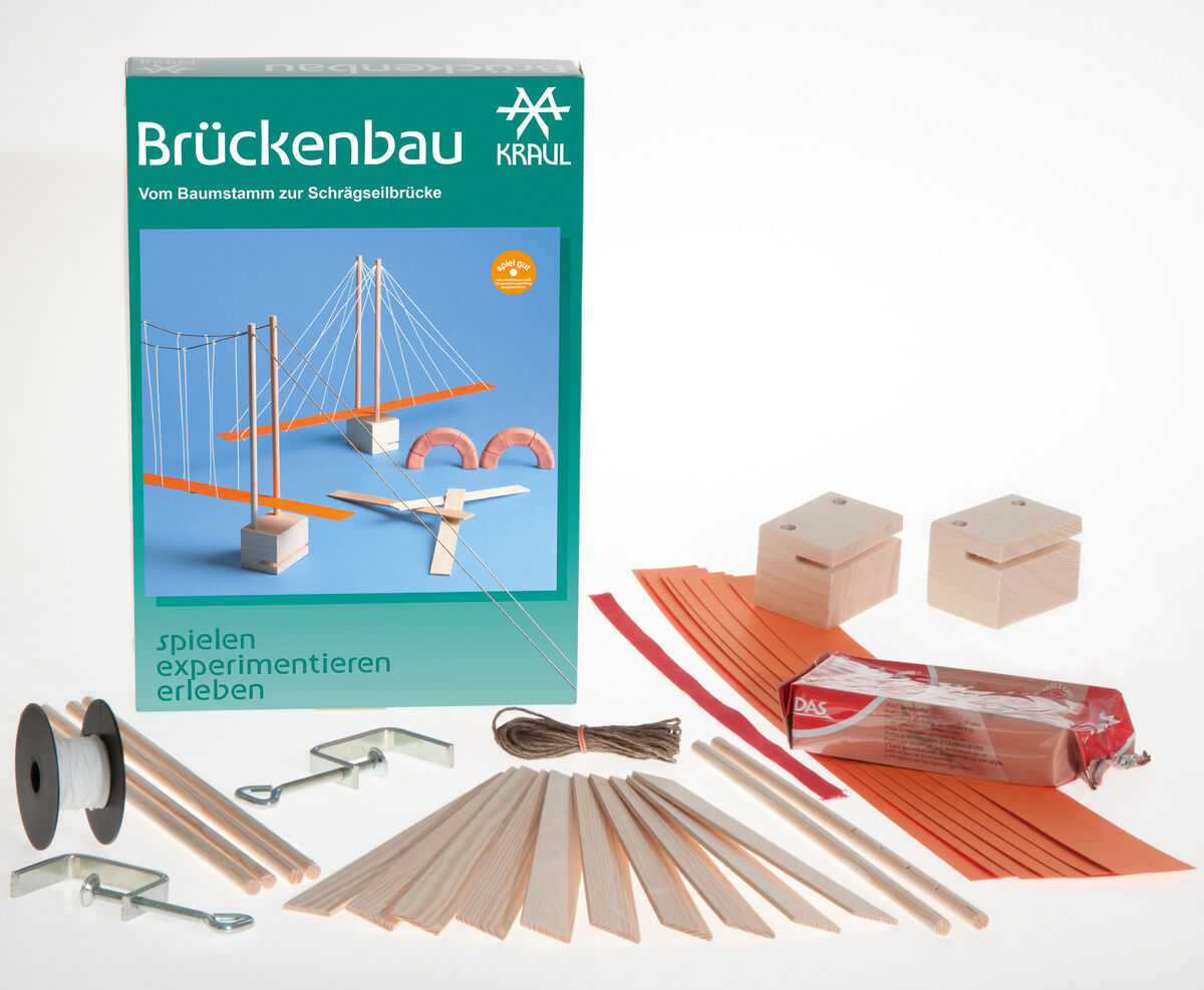 Foto eines Brückenbau-Bausatzes der Marke Kraul mit einer grünen Verpackung und verschiedenen Holz- und Bastelmaterialien wie Stäben, Schnur, Kartonteilen und Befestigungselementen. Der Bausatz ist für kreatives Experimentieren und das Nachbauen von Brücken geeignet.