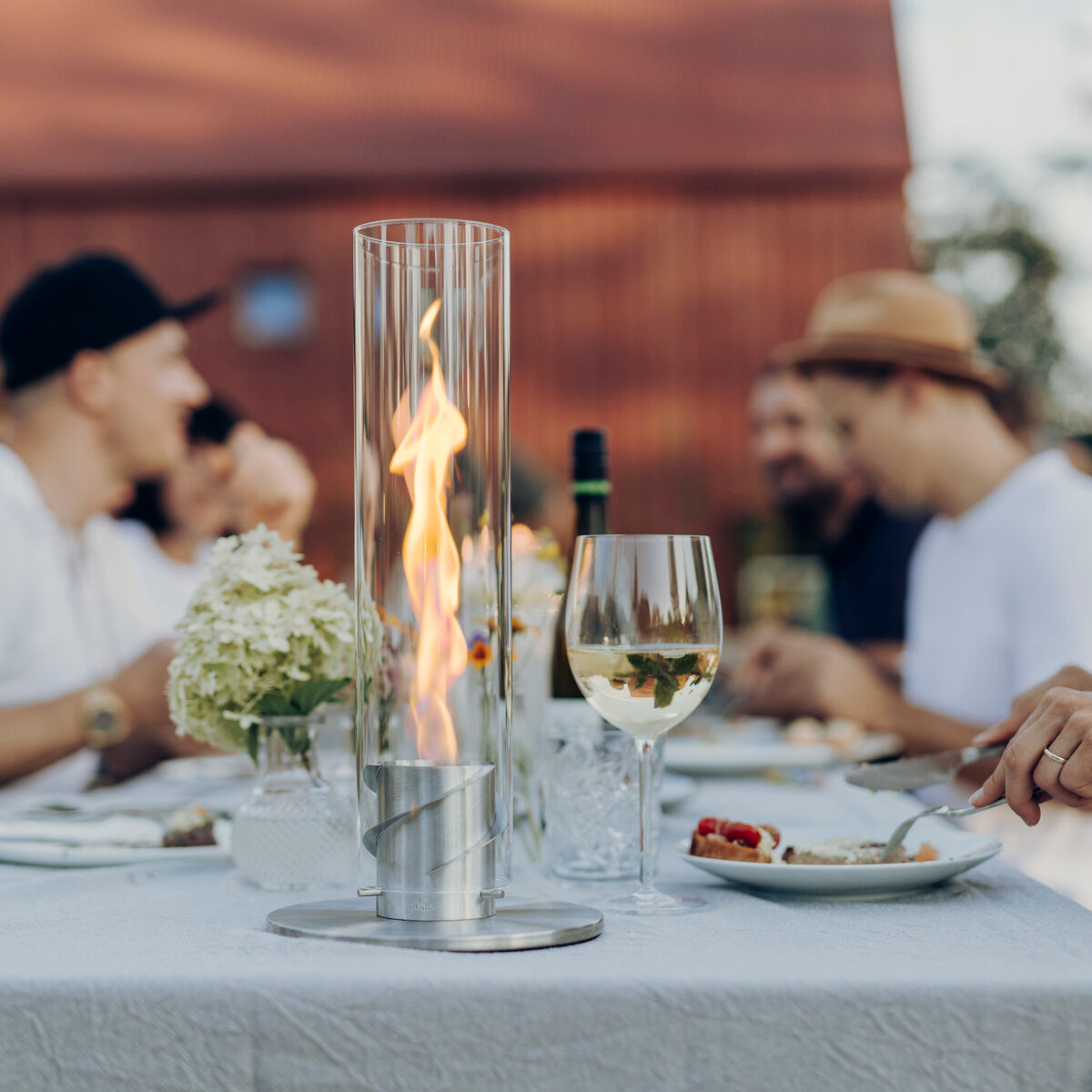 Menschen sitzen um einen Tisch, auf dem sich ein Tischfeuer SPIN 900 befindet. Im Hintergrund ist eine rote Scheune zu sehen. Der Tisch ist weiß gedeckt und es stehen Weingläser und Teller mit Essen darauf. Eine Frau im Vordergrund lächelt, während andere Menschen im Hintergrund miteinander sprechen.