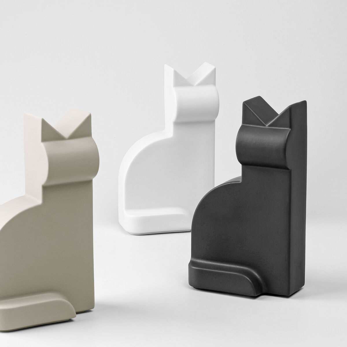 Das Bild zeigt drei Porzellanfiguren in Form von Katzen. Eine Figur ist schwarz, eine weiß und eine beige. Die Figuren haben ein minimalistisches und modernes Design.