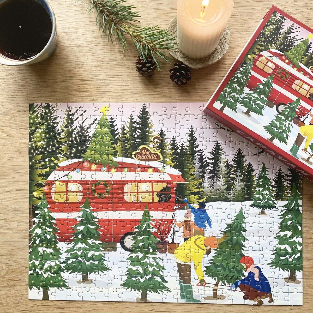 Das fertig zusammengesetzte 300-Teile-Puzzle „Caravan“ von ViSSEVASSE zeigt eine verschneite Winterlandschaft mit einem roten Wohnwagen und Menschen, die Weihnachtsbäume aussuchen. Daneben eine brennende Kerze, Tannenzweig, Tannenzapfen und eine Tasse Kaffee auf einem Holztisch.