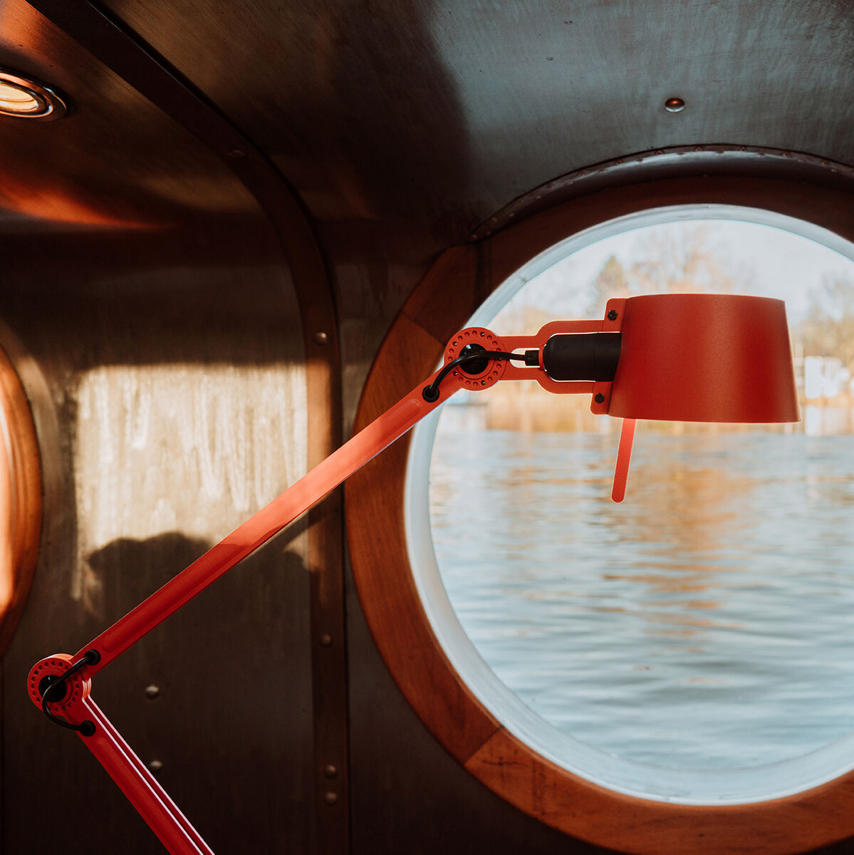 Eine rote, moderne Stehlampe mit zwei Armen befindet sich in einem gemütlichen Innenraum vor einem runden Fenster, das einen Blick auf eine friedliche Wasserlandschaft bietet.