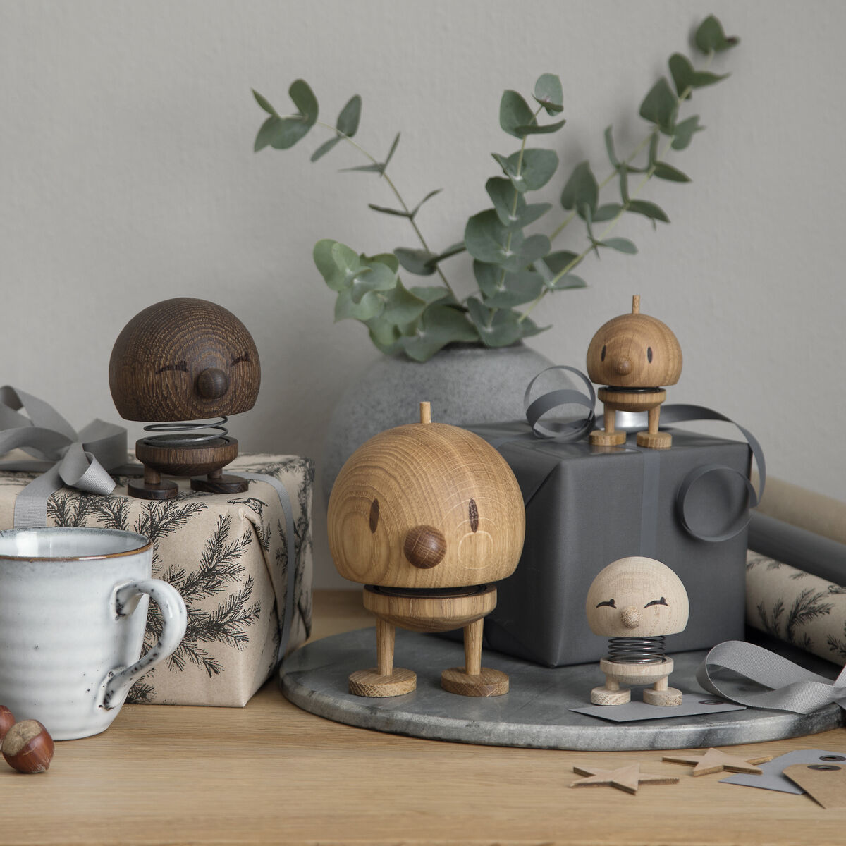 Vier Hoptimist Bumble Figuren aus Holz auf einem Tisch. Zwei Geschenkpakete, ein grauer Blumentopf mit Eukalyptus, eine weiße Tasse und Walnüsse sind ebenfalls zu sehen. Hintergrund ist eine graue Wand.