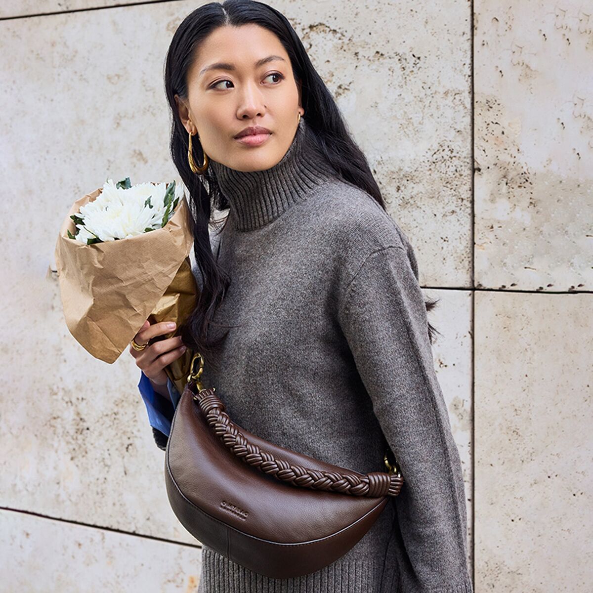 Eine Frau mit langen dunklen Haaren trägt eine schokoladenbraune Crossbody-Bag »Leo« von O My Bag aus offenporigem Leder sowie einen Strickpullover in Taupe. Sie hält einen Strauß weißer Blumen vor einer beigen Steinwand und blickt nach links.