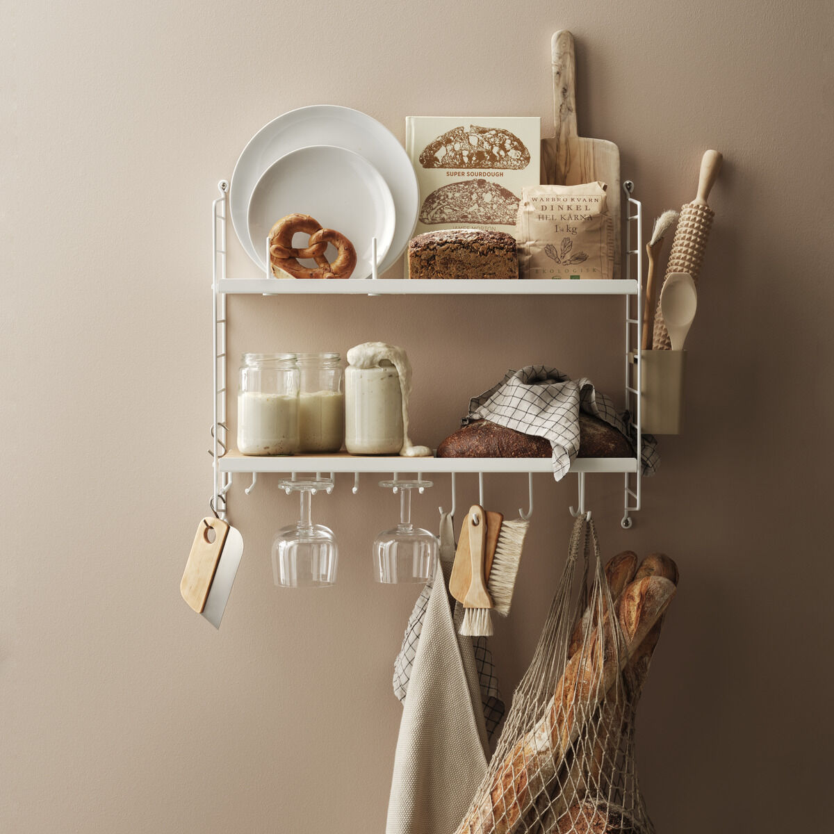 Das Bild zeigt ein weißes, modernes Küchenregal von String an einer beige gestrichenen Wand. Auf dem Regal stehen Schüsseln, Bücher, ein Holzbrett und Gläser. Zudem hängen Geschirrtücher und ein Netz mit Brot am Regal.