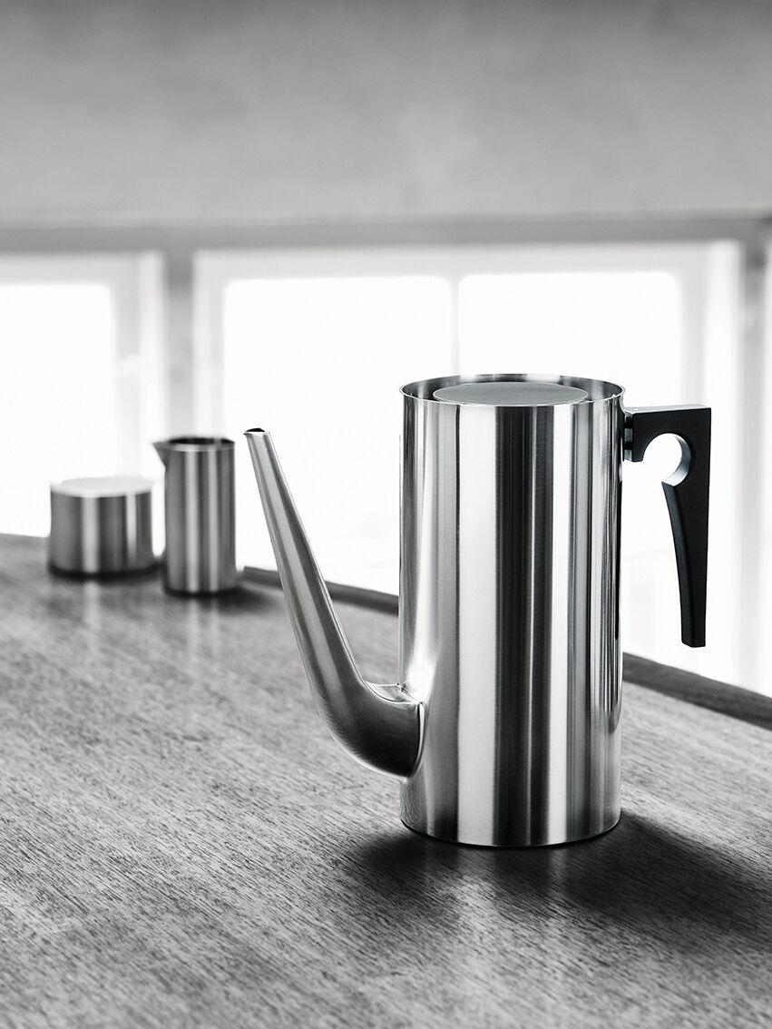 Eine stilvolle Kaffeekanne im modernen Design von Arne Jacobsen, hauptsächlich aus Edelstahl mit einem schwarzen Griff. Die Kanne steht auf einer Holztischplatte mit weiteren Edelstahlobjekten im Hintergrund. Das Design ist minimalistisch und zeitlos.