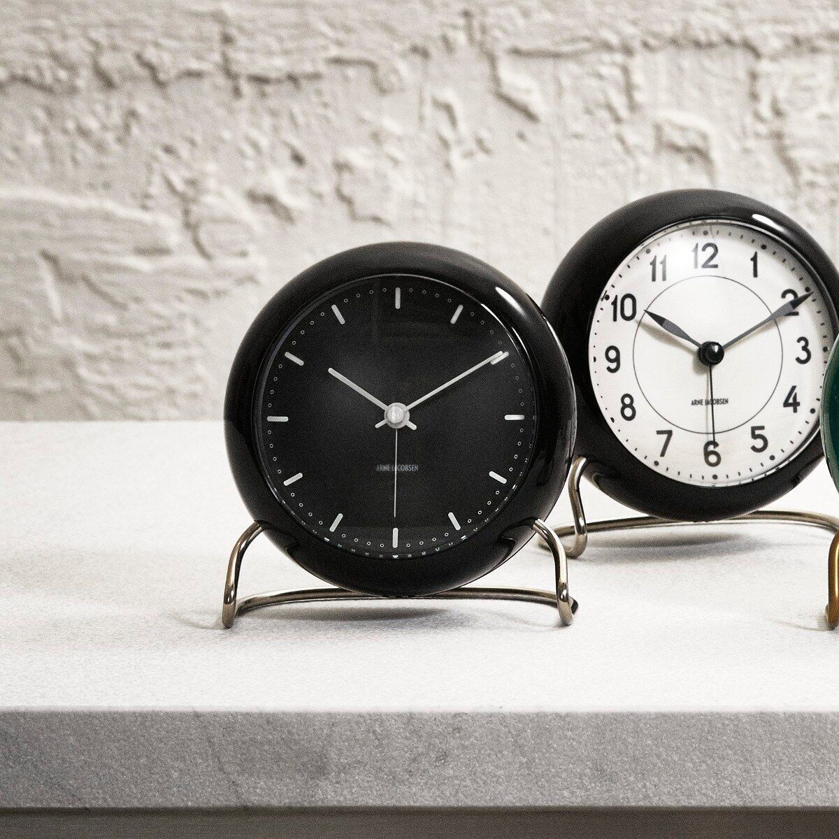 Zwei moderne Tischuhren »AJ City Hall« von Arne Jacobsen stehen auf einem hellen Tisch vor einer strukturierten, hellen Wand. Eine Uhr hat ein schwarzes Zifferblatt mit weißen Zeigern, die andere ein weißes Zifferblatt mit schwarzen Zahlen und Zeigern. Beide Uhren zeichnen sich durch ihr minimalistisches dänisches Design aus.