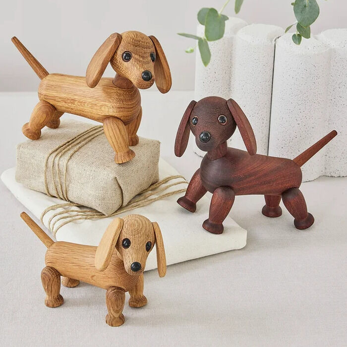 Drei Holzfiguren "Dackel Quick" von Spring Copenhagen stehen dekorativ auf einem hellen Tisch. Zwei Dackel sind aus hellem Holz, einer aus dunklem. Einer der hellen Dackel steht auf einem Geschenkpaket. Im Hintergrund sind grüne Eukalyptuszweige und weiße Rollen zu sehen.
