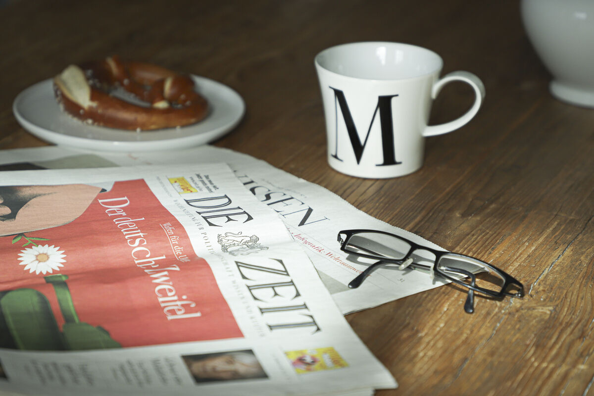 Eine weiße Frühstückstasse mit dem Buchstaben 'M' darauf steht auf einem Holztisch neben einer Zeitung 'Die Zeit', einer Brille und einer Brezel.