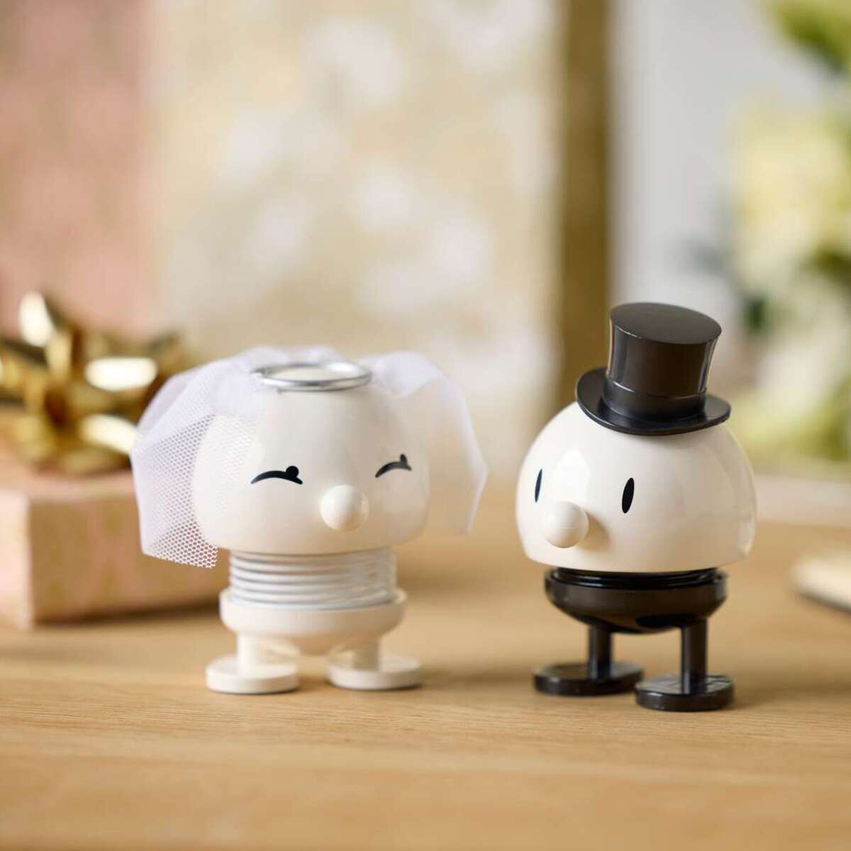 Zwei Hoptimist-Figuren als Braut und Bräutigam mit Schleier und Zylinder stehen auf hellem Holztisch. Im Hintergrund verschwommen weiße Blumen, ein Geschenk mit Goldschleife und ein aufgeschlagenes Buch. Szene wirkt festlich und freundlich dekoriert.