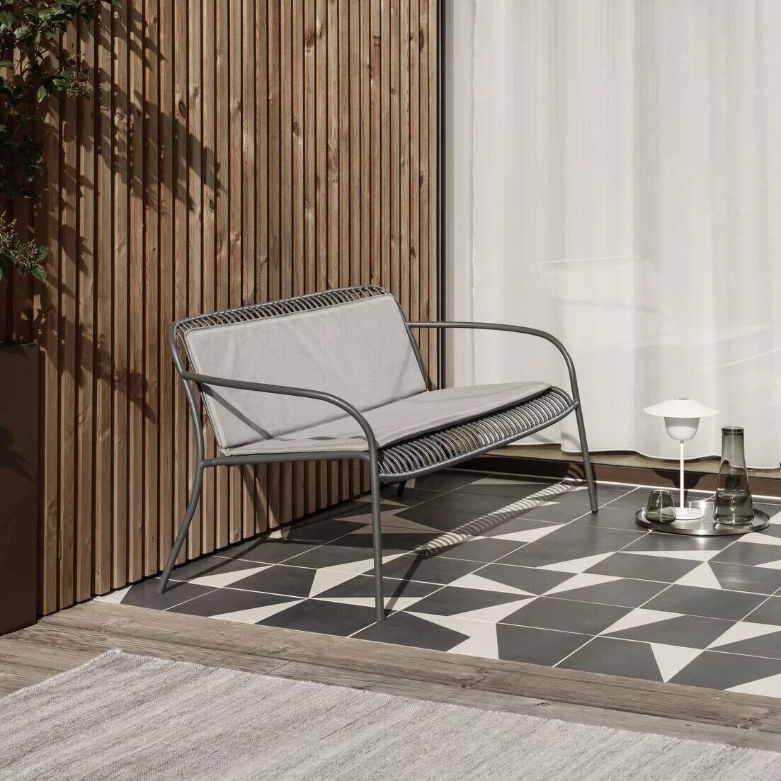 Eine stilvolle Scene auf einer Terrasse mit einer modernen grauen Bank, einem kleinen Tisch und einer mobilen LED-Tischleuchte »ANI«. Im Hintergrund sind Holzwände und ein großer Glasvorhang zu sehen.