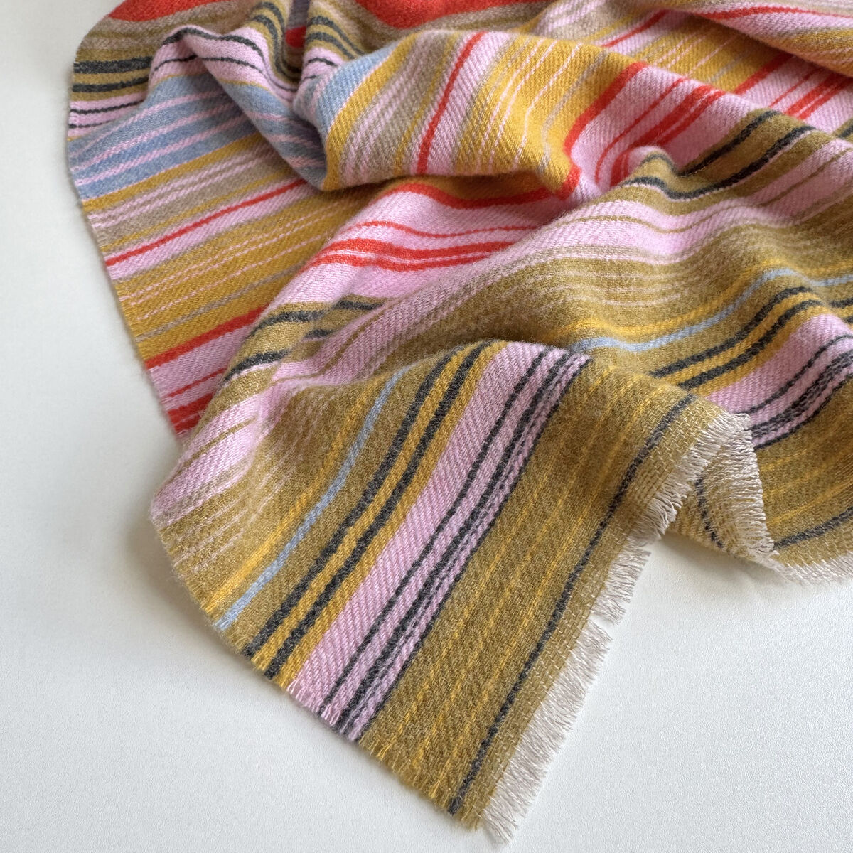 Nahaufnahme des locker drapierten Wollschals »Ariana« von Eagle Products auf hellem Untergrund. Der weiche Merino-Schal zeigt unregelmäßige, feine Streifen in Senfgelb, Rot, Rosa, Hellblau, Grau, Beige und Schwarz. Im Vordergrund liegt ein breites Ende mit kurzen, hellen Fransen; die Falten betonen die textile Struktur.