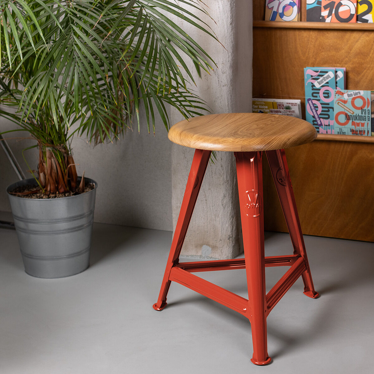 Ein Hocker aus der ZEIT-Sonderedition Rowac-Schemel »Red Brick« ist in einem Raum neben einer Pflanze und einem Regal zu sehen. Der Hocker hat eine Holzsitzfläche und rote Metallbeine.