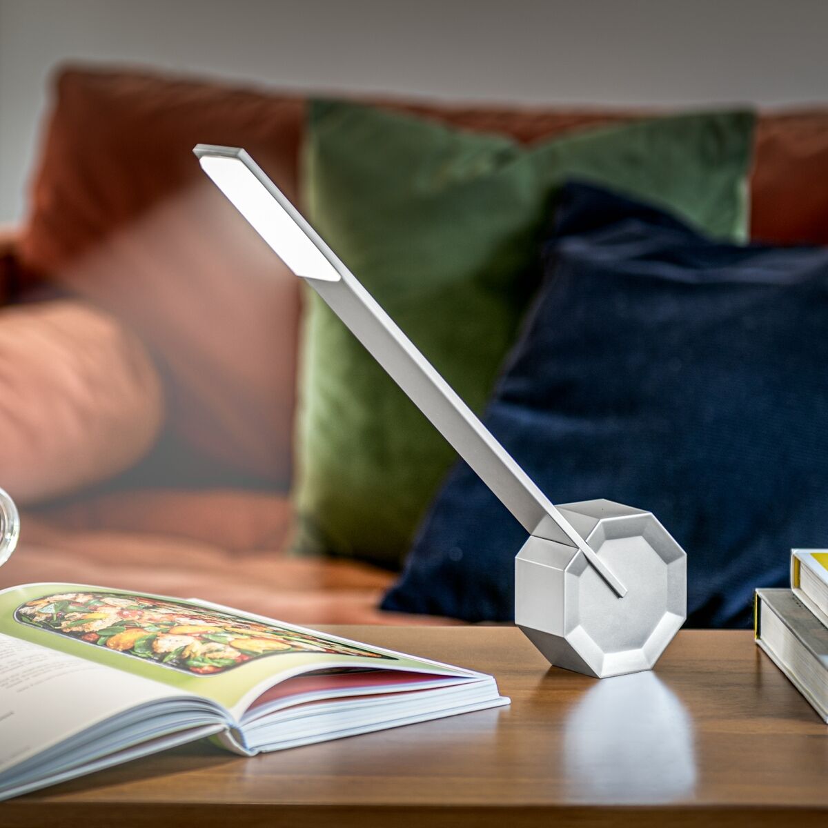 Eine moderne Tischlampe mit achtkantigem Design und schlanken LED-Leuchten, platziert auf einem eleganten Holztisch. Im Hintergrund befinden sich ein Sofa mit farbigen Kissen und ein aufgeschlagenes Buch.