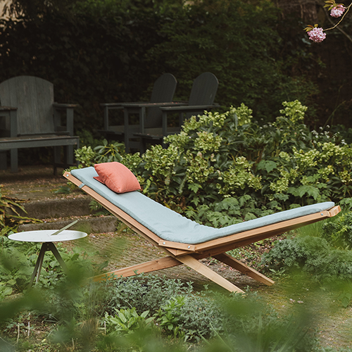 Liegestuhl mit hellblauer Polsterung und rotem Kissen in einem grünen Garten, daneben ein kleiner runder Beistelltisch, im Hintergrund zwei dunkle Gartenstühle auf einer Treppe.