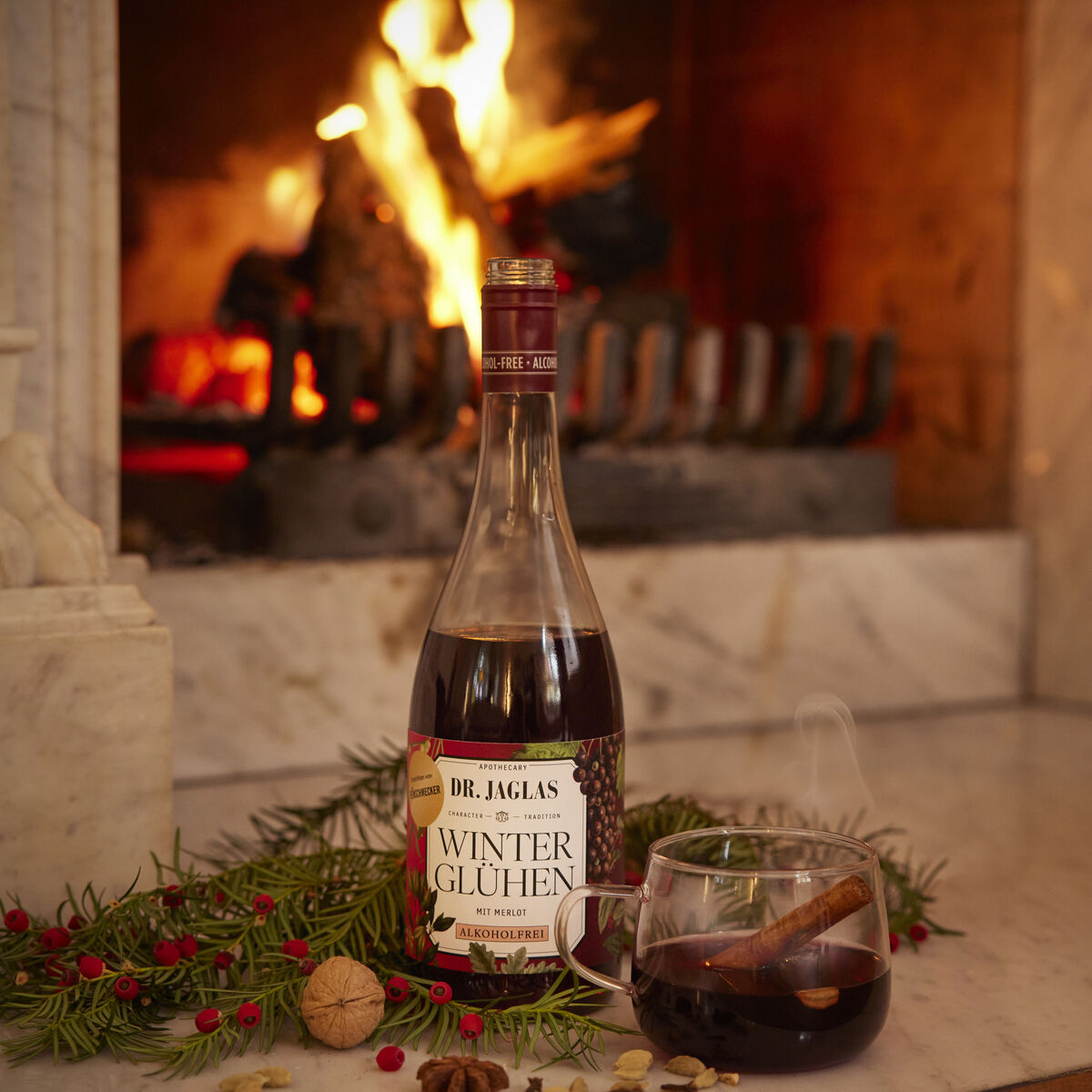 Eine Flasche Dr. Jaglas Winterglühen Merlot alkoholfreier Winzerglühwein steht auf hellem Marmor vor einem prasselnden Kaminfeuer. Daneben liegen weihnachtliche Deko wie Tannenzweige, rote Beeren, eine Walnuss, Sternanis und ein Glas Glühwein mit Zimtstange.
