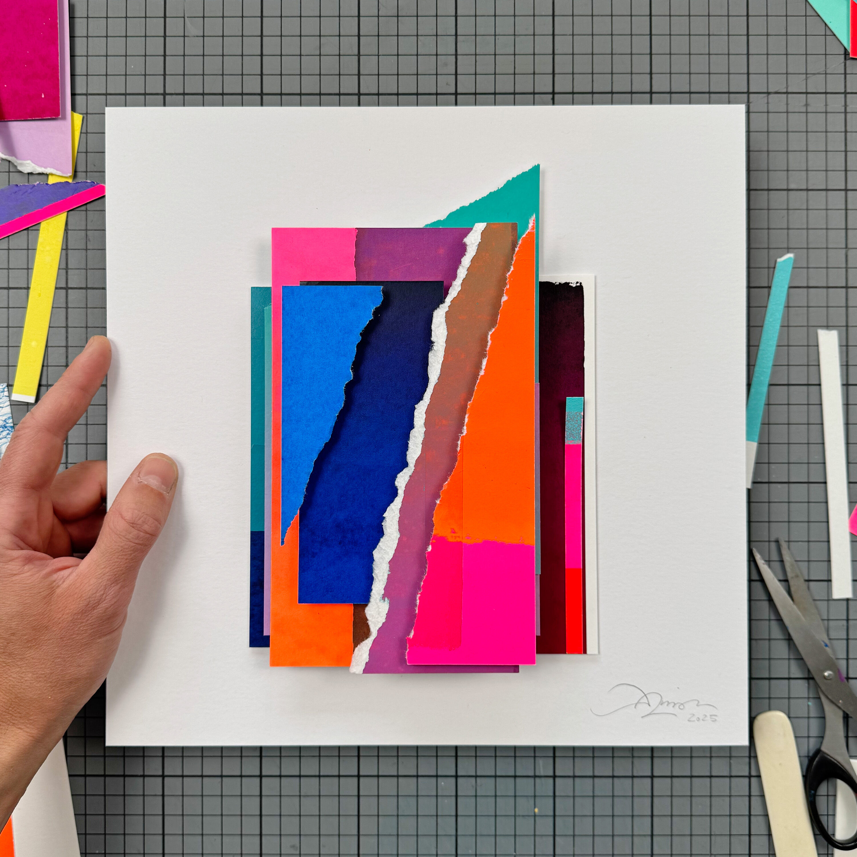 Handgefertigte Collage aus farbigen, gerissenen Papierstreifen in leuchtenden Pink-, Orange-, Blau- und Rottönen, kunstvoll auf weißem Karton arrangiert. Eine Hand hält das Kunstwerk, Bastelmaterialien liegen daneben. Moderne, abstrakte Siebdruck-Arbeit, ideal als Wohnungsdekoration oder Geschenk.