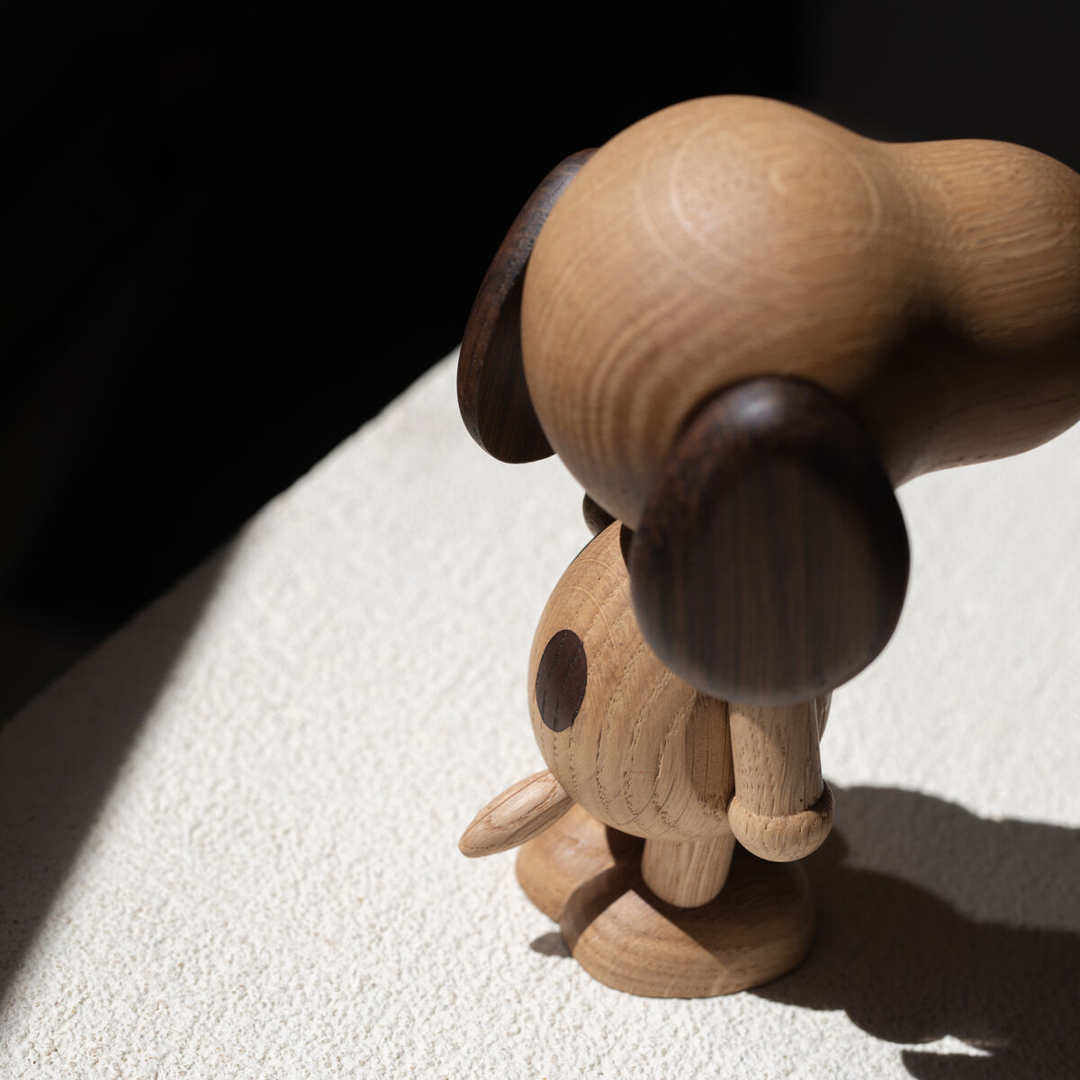 Eine Holzfigur von Snoopy steht auf einer weißen, strukturierten Oberfläche im Sonnenlicht. Die Figur ist aus hellem Naturholz mit dunkelbraunen Details gefertigt.