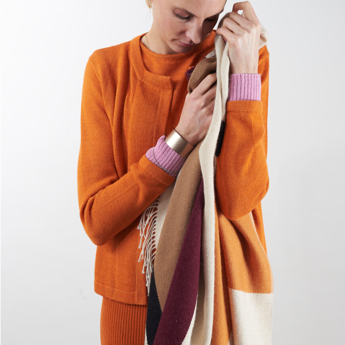 Eine Person trägt ein oranges Cape und hält ein mehrfarbiges Tuch. Das Cape besteht aus Alpakawolle und die Farben des Tuchs umfassen rot, beige, taupe und weiß.