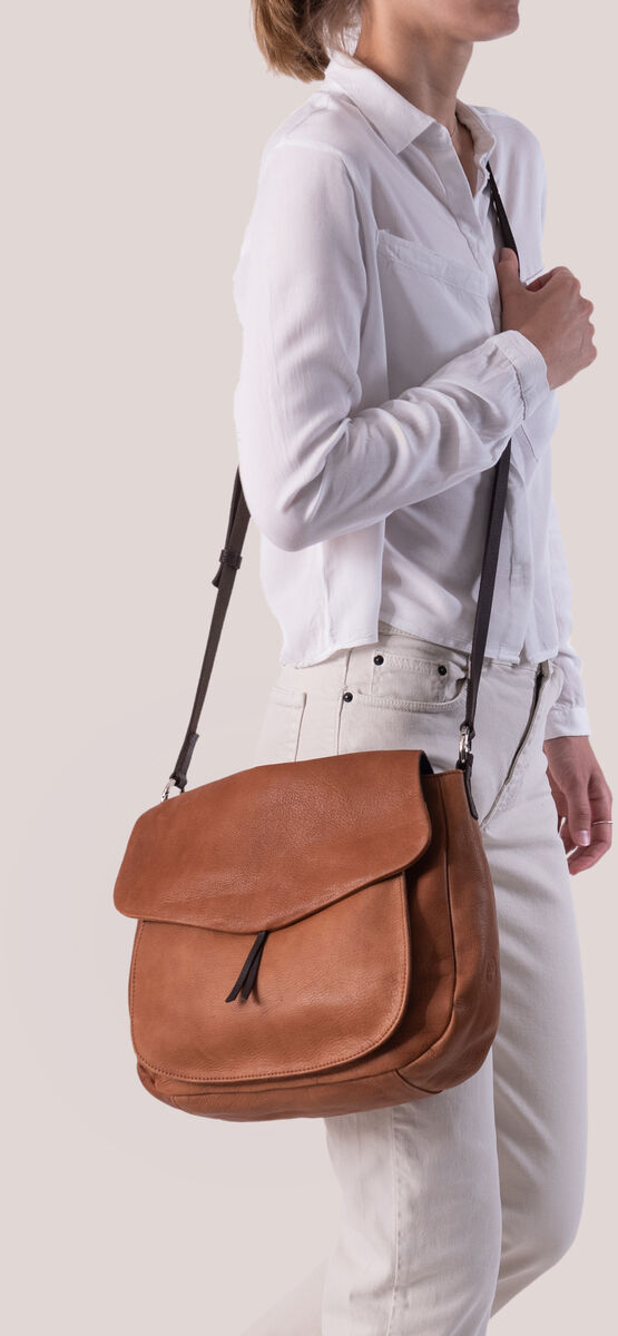 Eine Frau trägt eine braune Leder-Umhängetasche (»Slingbag«). Sie trägt ein weißes Hemd und eine helle Hose. Der Hintergrund ist schlicht und einfarbig gehalten.
