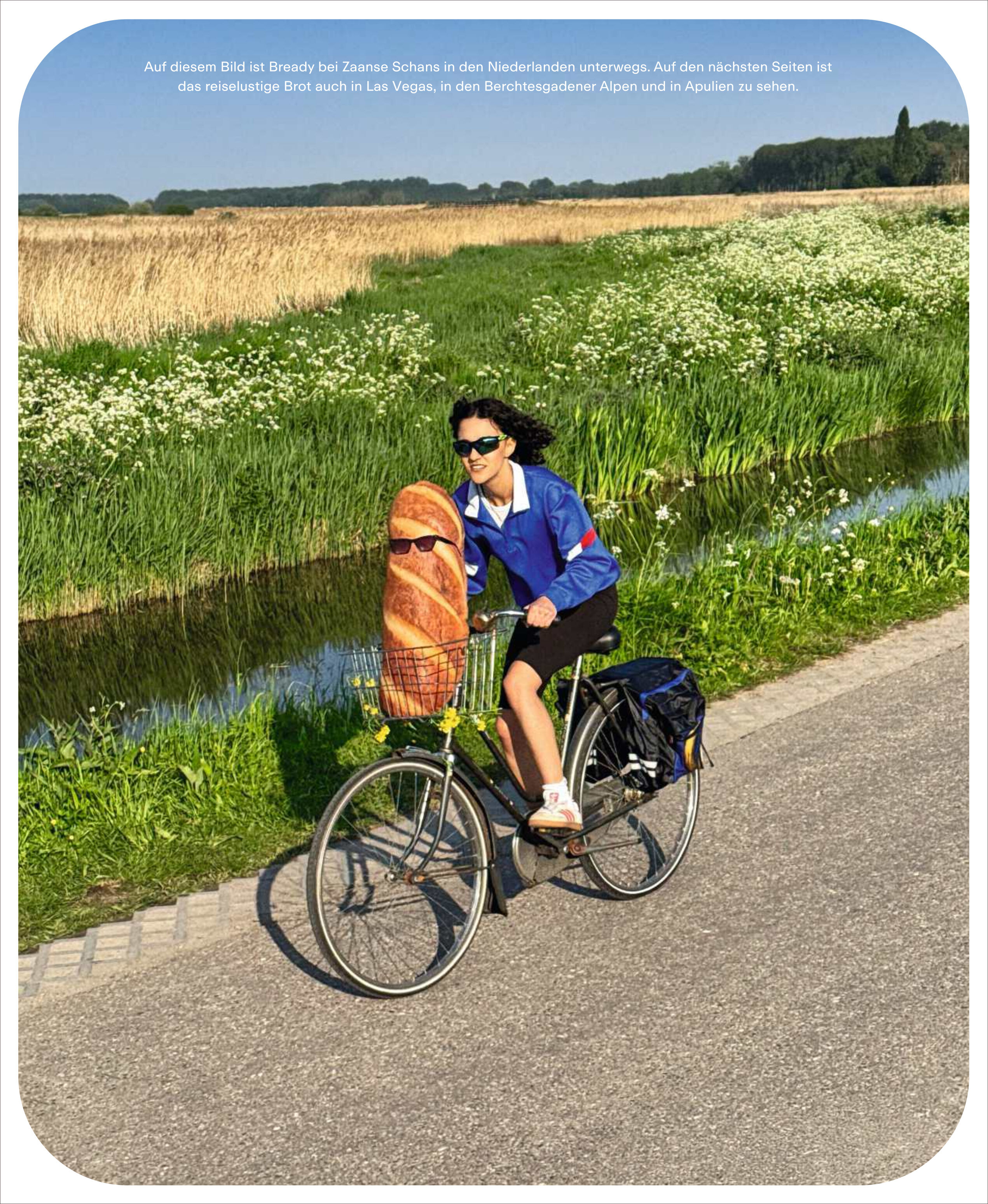 Eine junge Frau fährt lachend auf einem Fahrrad einen asphaltierten Weg entlang, vorbei an einer grünen Wiese mit hohem Gras und einem Wassergraben. In ihrem Fahrradkorb liegt ein großes Baguette. Die Szene ist bei sonnigem Wetter aufgenommen. Die gesamte Atmosphäre vermittelt Sommer, Freiheit und Reiselust.