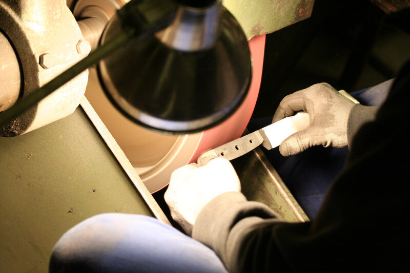 Eine Person mit Handschuhen hält ein Kräutermesser »Shark« und schärft es an einer Schleifmaschine unter einer Lampe.