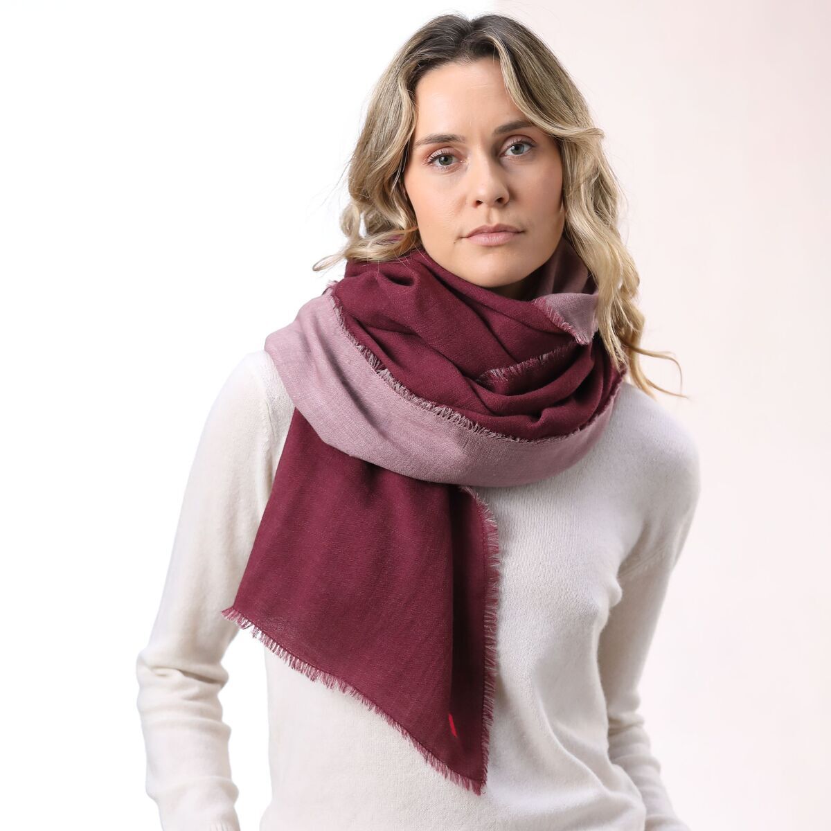 Frau trägt einen kuscheligen Doubleface-Wollschal mit Vorderseite in Bordeauxrot und Rückseite in Rosé über einem cremefarbenen Pullover und einer hellen Hose vor neutralem Hintergrund. Der Schal ist stilvoll und elegant drapiert und verleiht dem winterlichen Outfit eine moderne Note.