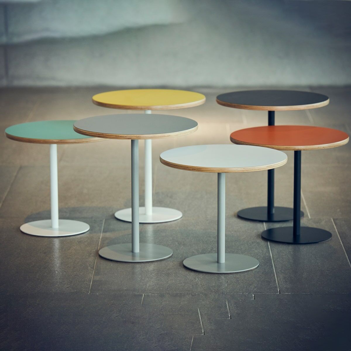 Eine Gruppe von sechs runden Beistelltischen des Modells »Panama« in verschiedenen Farben (weiß, grau, schwarz, hellgrün, senfgelb, und terracotta) mit Metallfuß auf einem grauen Boden.