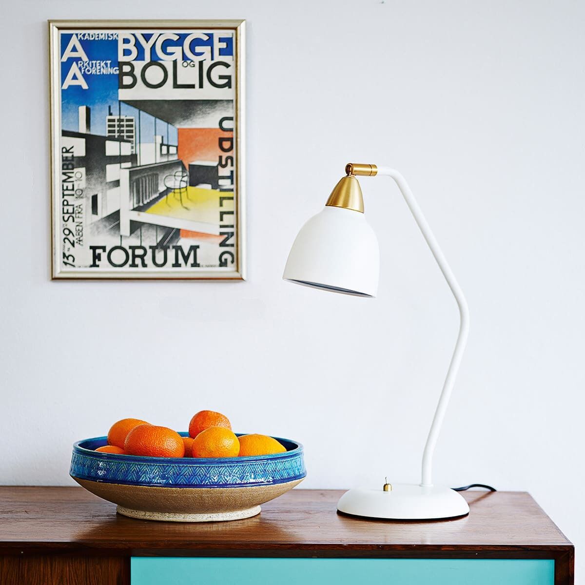 Ein modernes Zimmer mit einer Tischlampe namens 'Urban Whisper White', die auf einem Holzschrank mit einer Schublade steht. Daneben eine Schale mit Orangen und im Hintergrund ein Kunstposter an der Wand.