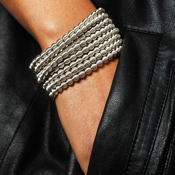Mehrreihiges silbernes Perlenarmband namens Chapelet wird am Handgelenk einer Person getragen, die einen schwarzen Lederärmel trägt. Das Armband besteht aus mehreren Perlenreihen, die eng aneinanderliegen und einen eleganten, modernen Look erzeugen.