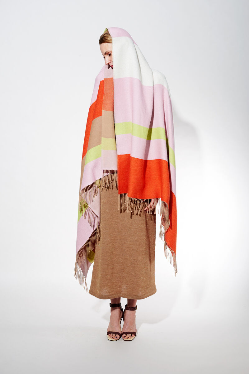 Eine Frau trägt ein Cape namens 'Blossom' aus Alpaka-Wolle. Das Cape hat ein Streifenmuster in den Farben Orange, Rosa, Weiß, Beige und Grün. Es hat Fransen an den Enden und wirkt sehr weich und gemütlich.