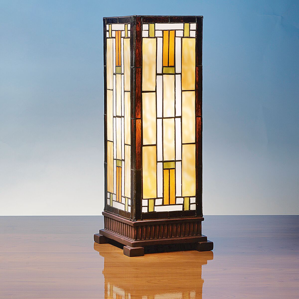 Rechteckige Tischleuchte »Carré« im Stil von Louis C. Tiffany, gefertigt aus mehrfarbigem Tiffany-Art-Glas mit geometrischem Muster in Bernstein-, Creme- und Brauntönen, auf einem kunstvoll verzierten dunklen Kunstguss-Sockel. Die Leuchte steht auf einer glänzenden Holzoberfläche vor blauem Hintergrund.