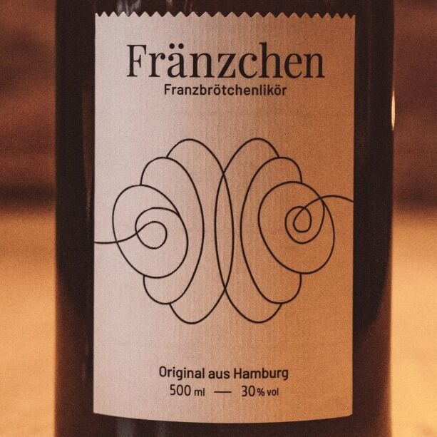 Eine stilvolle Flasche des Likörs 'Fränzchen' mit einem kunstvollen Etikett und Korkverschluss. Die Flasche steht auf einem Holztisch vor einem unscharfen, warmen Hintergrund.