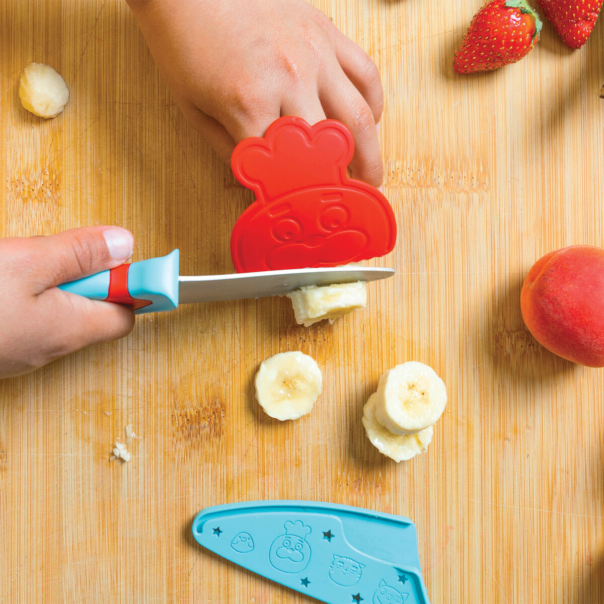 Ein kindgerechtes Messer mit einem fröhlichen Design schneidet Bananenscheiben auf einem Holzbrett. Ein Kind hält das sichere, ergonomisch gestaltete Messer, während auf dem Brett auch Erdbeeren und ein Pfirsich zu sehen sind.