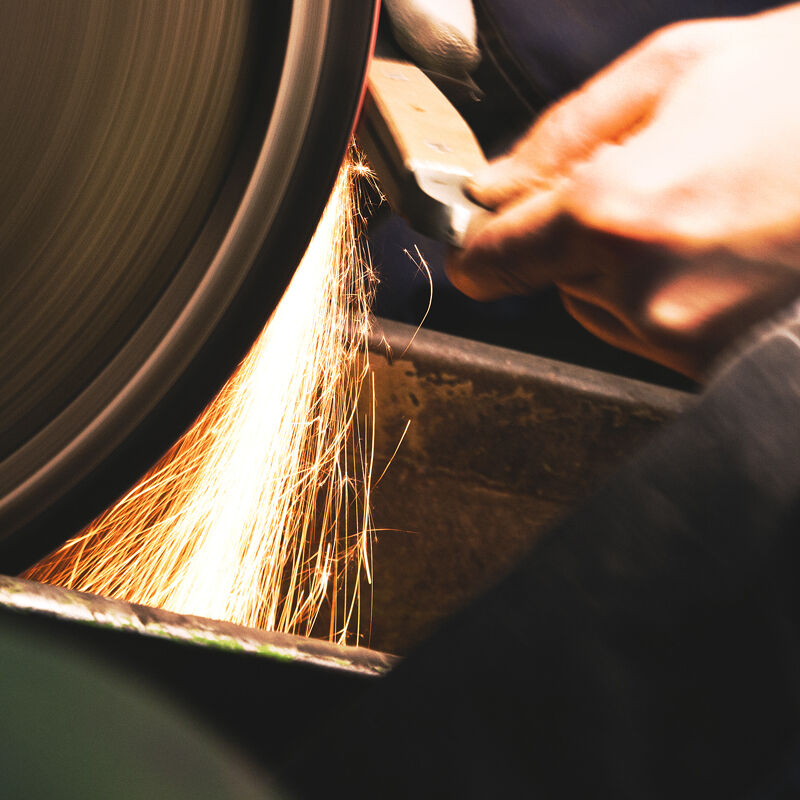 Ein Käsemesser mit Olivenholzgriff wird gerade an einem Schleifstein geschärft, wobei Funken sprühen. Das Bild zeigt ein handgefertigtes, qualitativ hochwertiges Küchenutensil in der Entstehung.