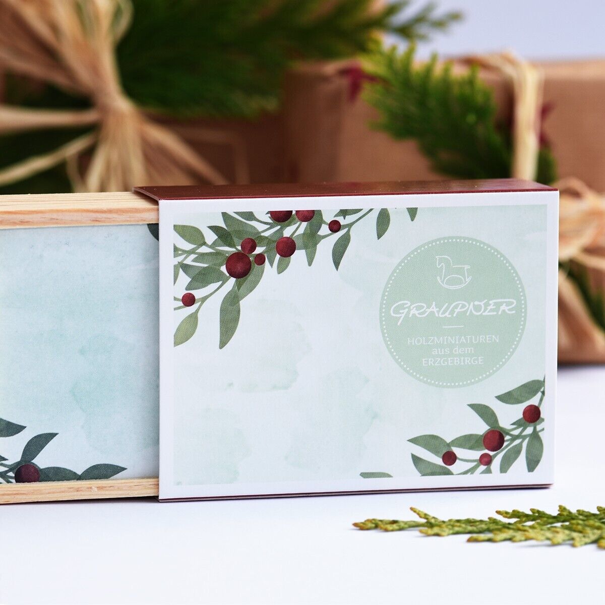 Eine dekorative Box mit weihnachtlichen Motiven und einem Etikett, das die Aufschrift 'Glückwunschkästchen »Krippe«' trägt. Im Hintergrund sind grüne Zweige und verpackte Geschenke zu sehen.
