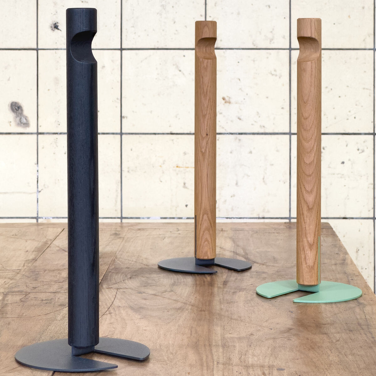 Das Bild zeigt drei moderne Küchenrollenhalter »GIVE« der Marke finbeaux von Christoph Schoen, aus Holz mit metallenen, unterschiedlich farbigen Fußplatten in Schwarz, Grau und Mintgrün, auf einer Holzplatte vor einer gefliesten Wand stehend.